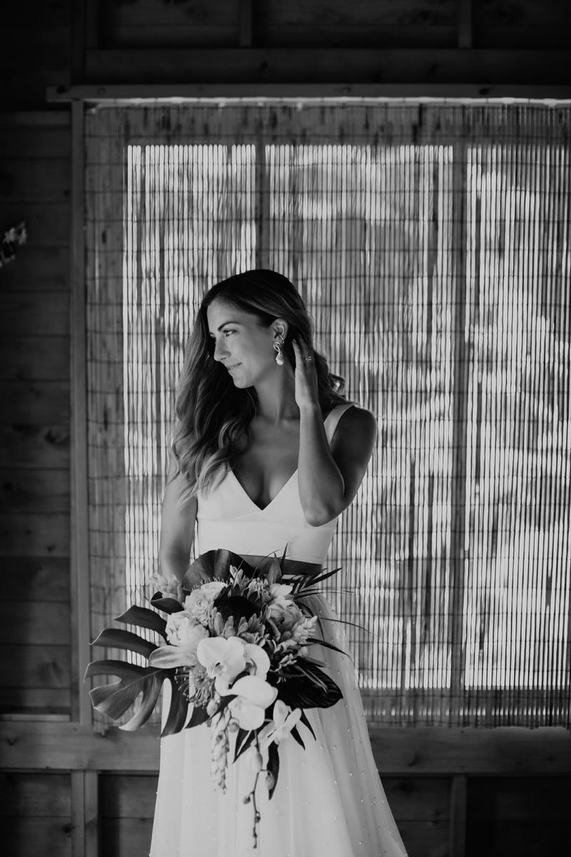 B&W photo of KWH real bride Jana standing in a wooden room with her native Hawaiian bouquet from Grace Flowers. She wears the v-neck Erin bodice paired with the aline Erin skirt.