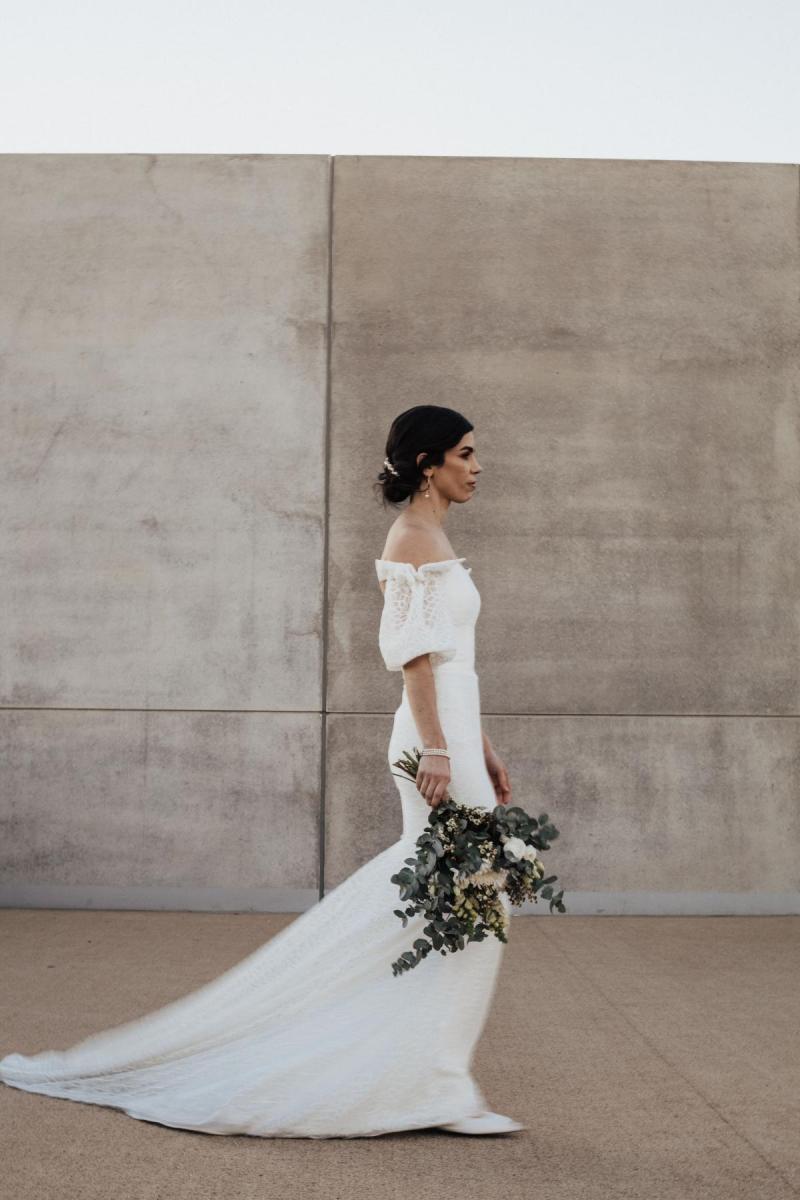 KWH real bride Tess walking by a cement wall in her fairytale Vivienne wedding dress with off the sholder puff sleeves.