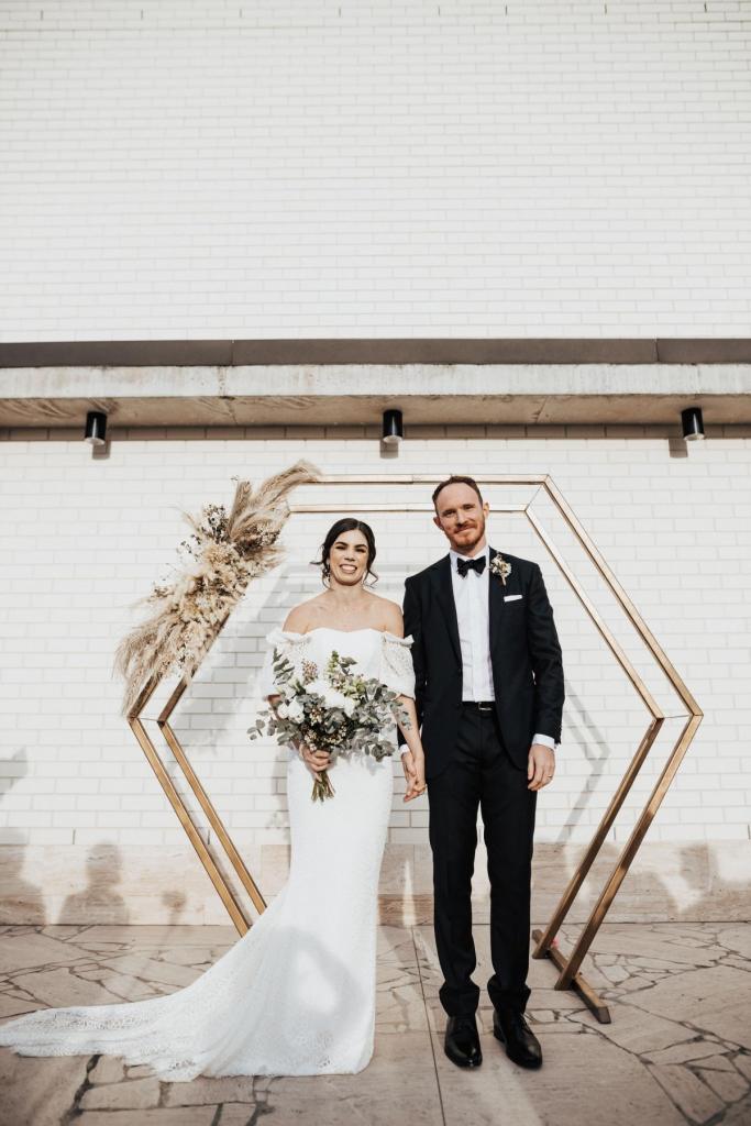 KWH real bride Tess standing at their hexagon gold alter with Liam. She dons the fitted Vivienne wedding dress with all over modern lace.