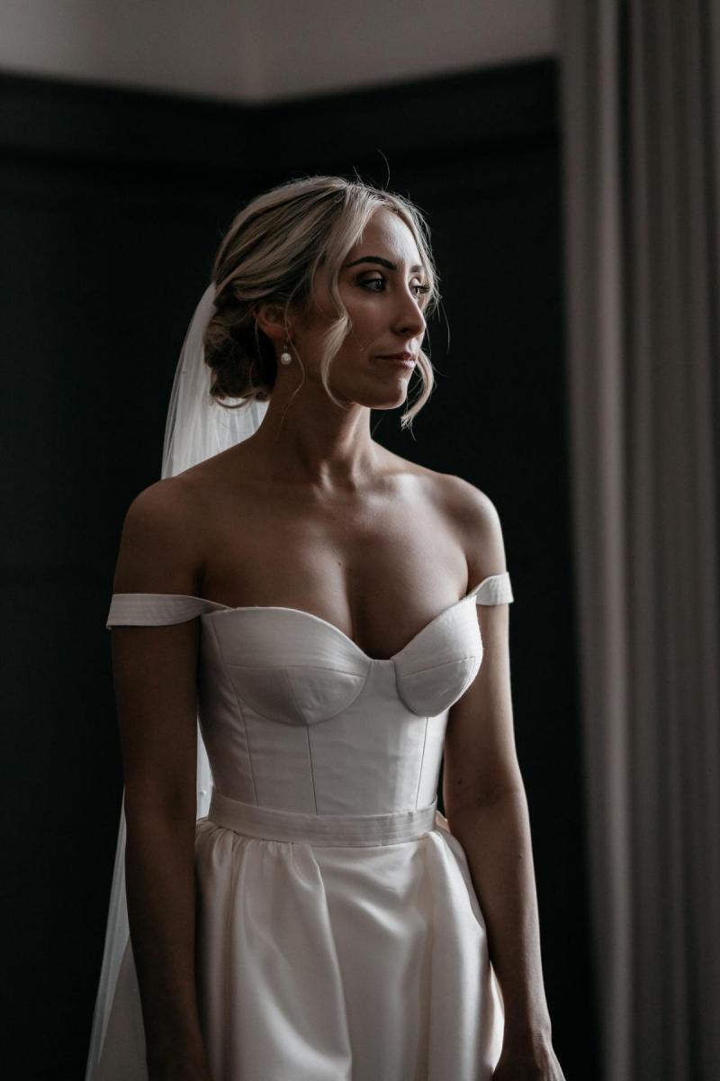 KWH real bride Genevieve stands in the glow of the window in her Blake Camille wedding dress; a modern aline gown with off-the-shoulder straps.