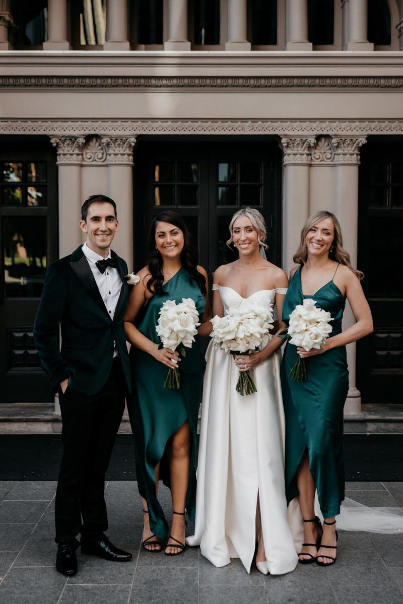 KWH real bride Genevieve stands outside with her bridal party who wear silk dark green dresses. She wears the timeless Blake Camille wedding dress.