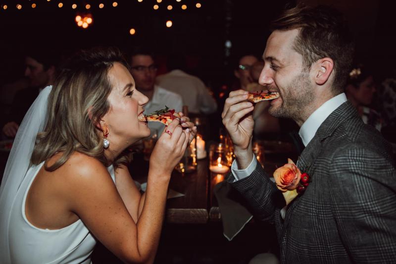 KWH real bride Casey and her new husband eat slices of pizza as she wears her ivory Bardot jumpsuit with wide leg pant and side cutaways.