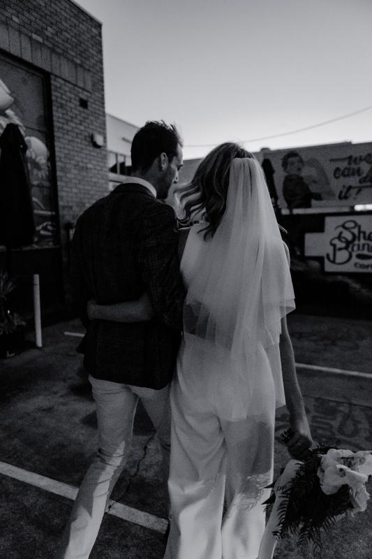 B&W image of Real bride Casey and her husband walking down a street. She wears the minimalist bridal jumpsuit, Bardot.