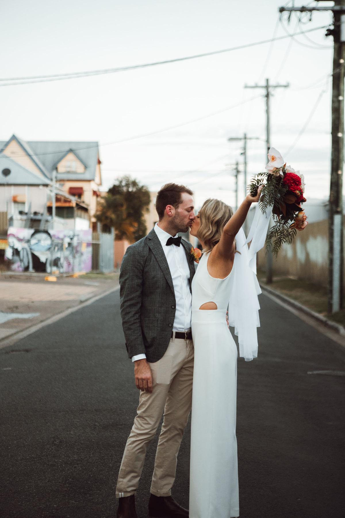 KWH real bride Casey and Pat smooch in the street. She wears the Bardot bridal jumsuit, a staight-leg modern jumpsuit.