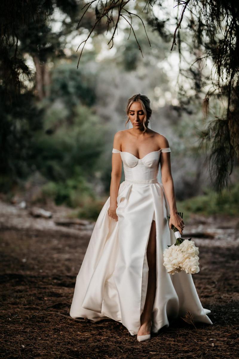 KWH real bride Genevieve walking through the woods in her Blake Camille wedding dress; a rouged a-line gown with bustier bodic and off the shoulder strap.