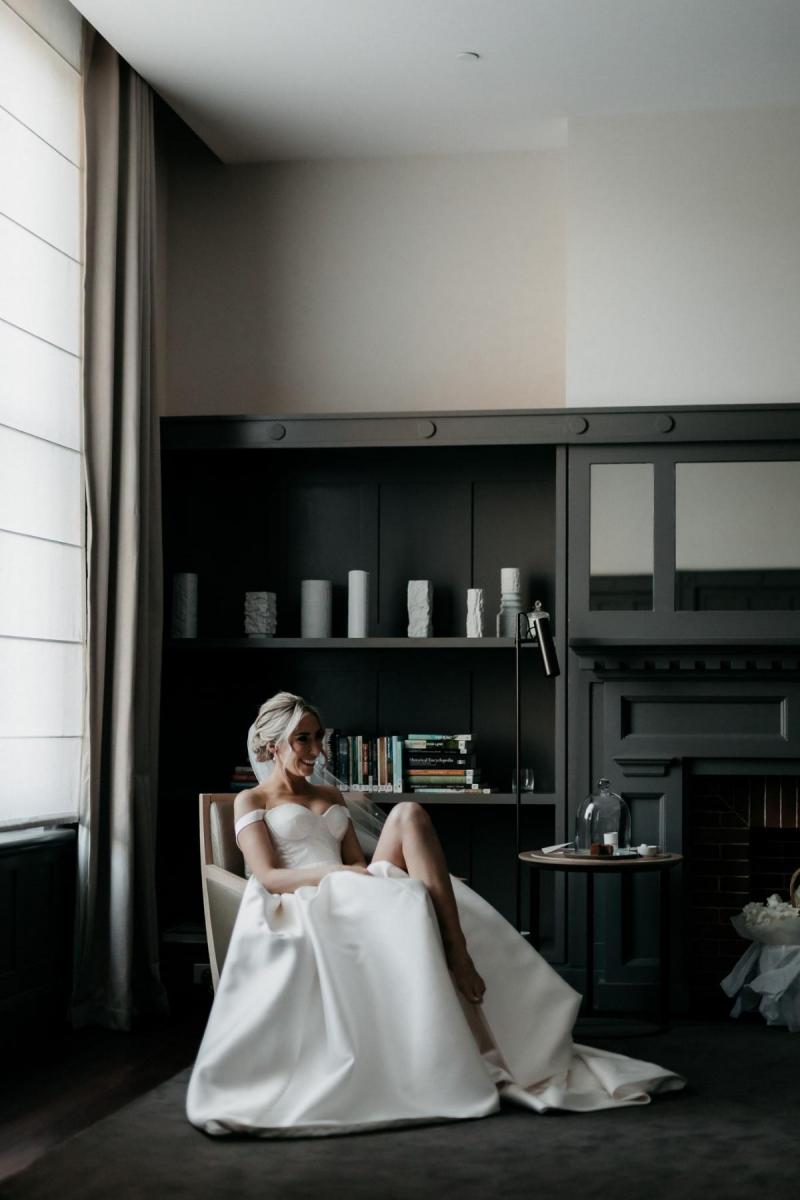 KWH real bride Genevieve sitting in a chair at the hotel. She wears the Blake Camlle modern a-line wedding dress with bustier bodice and off the shoulder strap