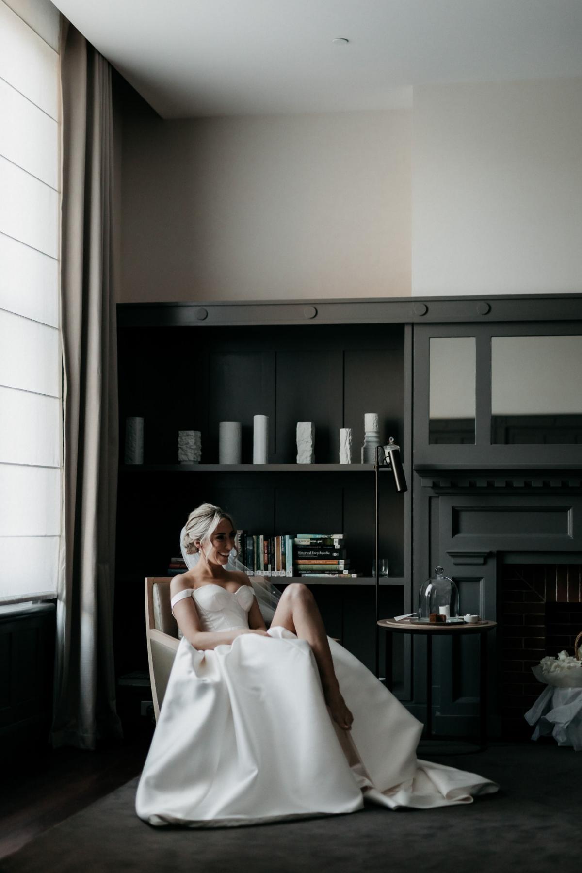 KWH real bride Genevieve sitting in a chair at the hotel. She wears the Blake Camlle modern a-line wedding dress with bustier bodice and off the shoulder strap