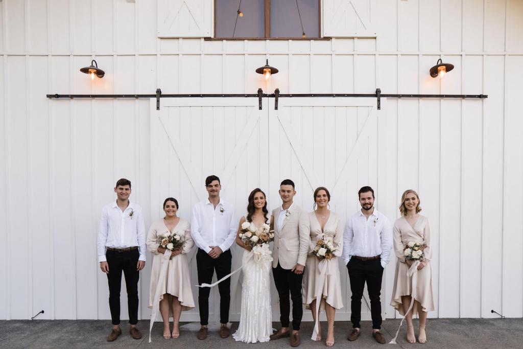 KWH real bride Georgie stands with her wedding party in front of the Summergrove Estate barn doors. She wears the Fontanne gown, a fit and flare beaded wedding dress.