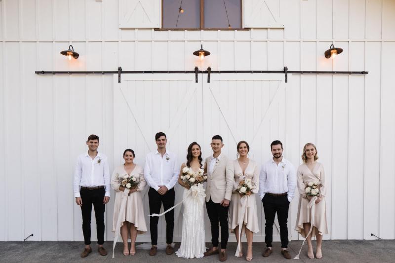 KWH real bride Georgie stands with her wedding party in front of the Summergrove Estate barn doors. She wears the Fontanne gown, a fit and flare beaded wedding dress.