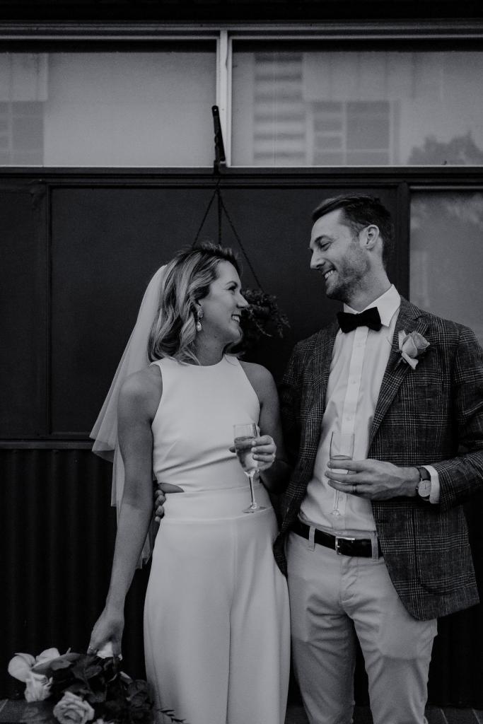B&W image of KWH real bride Casey and Pat holding their celebratory drinks. She wears the modern Bardot jumpsuit, a high neck staight-cut leg bridal jumpsuit