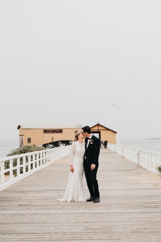 KWH real bride Nicole and Chris walk down the beachside pier. She wears the vintage inspired Cassie wedding dress with long sleeves and keyhole back.
