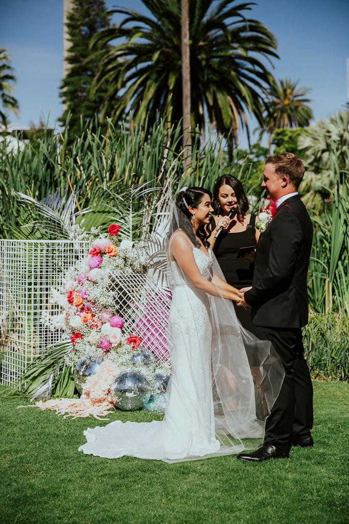 KWH real bride Jaz and Chris standing at their colourful tropical alter. She wears the Darcy gown, a modern fit and flare sequin wedding dress.