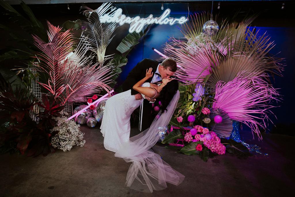 KWH Real bride Jaz being dipped as they dance by Chris in front of neon light decor. She wears the modern Darcy gown, a beaded v-neck wedding dress.