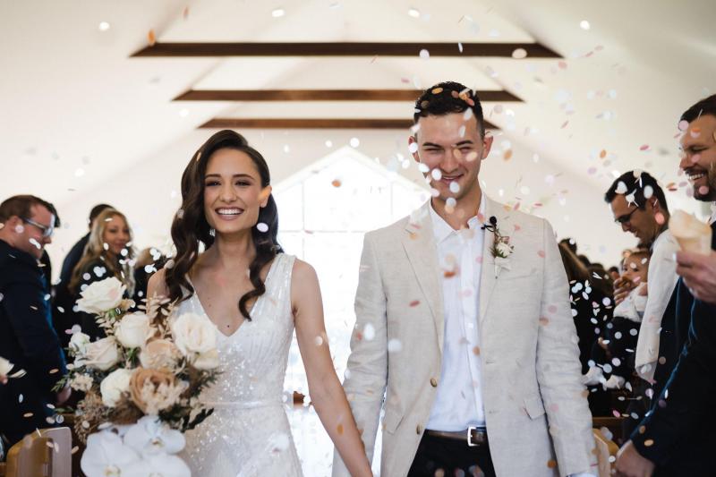 KWH real bride Georgie and new husband Mikey walk down the aisle with confetti thrown. She wears the Fontanne gown, a v-neck beaded wedding dress.