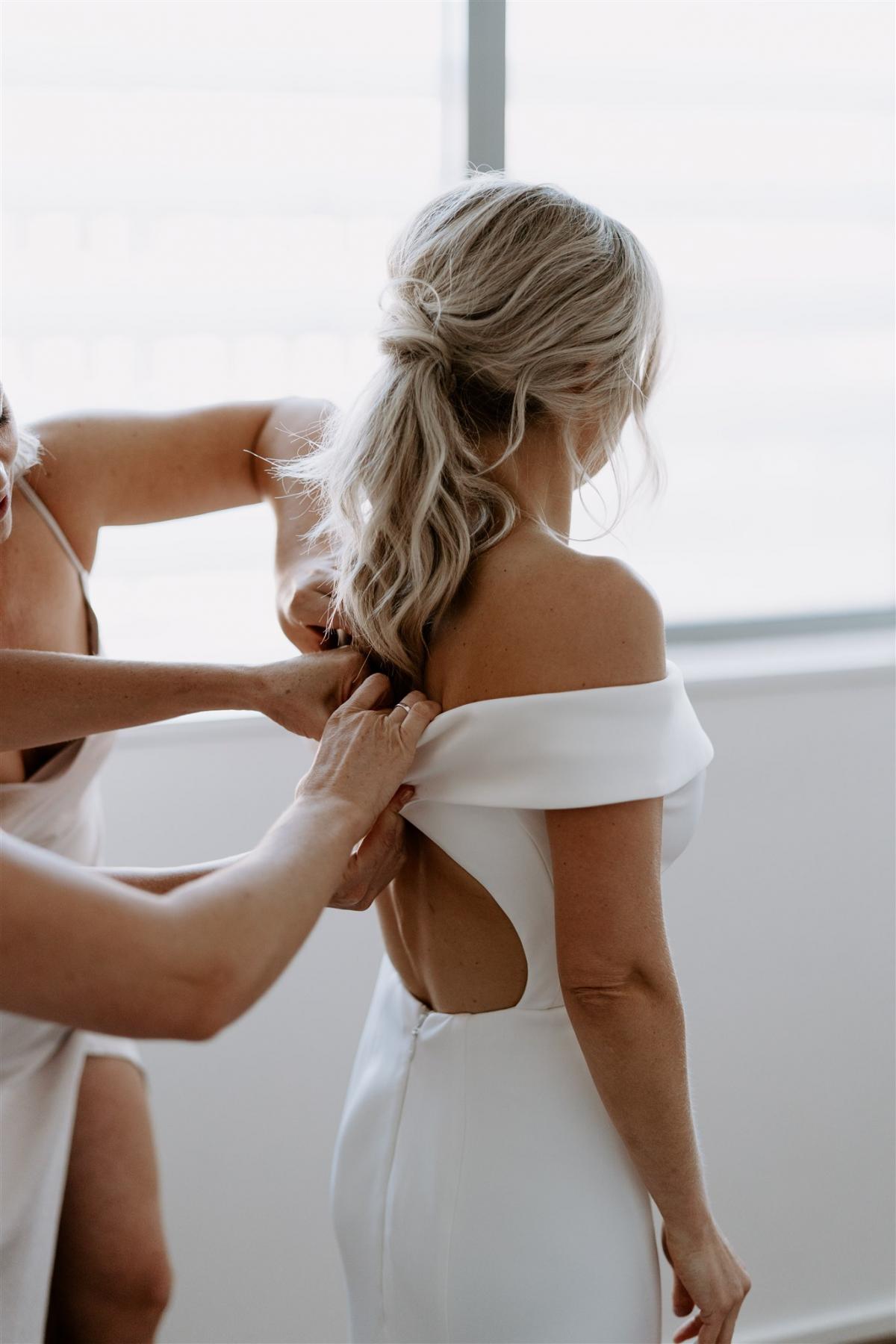 KWH real bride Lauren has her bridesmaid do up her satin buttons on her Lauren gown; a timeless ivory wedding dress with off the shoulder sleeve.