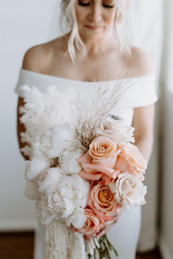 KWH real bride Shelley poses with her incredible peachy florals by Poppy's Flowers. She wears the classic Lauren wedding dress with off the shoulder sleeve and fit and flare silhouette.