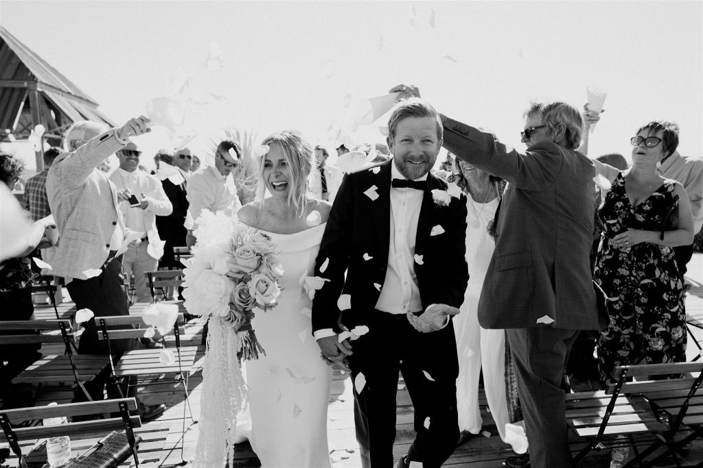 B&W image of KWH real bride Shelley and Jack walking down the aisle as petals are thrown. She wears the timeless Lauren wedding dress with fit and flare silhouette.