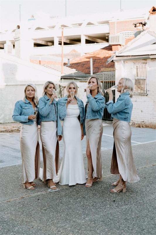 KWH real bride Shelley stands with her bridesmaids who are wearing denim jackets. She wears the sophisticated Lauren wedding dress with off the shoulder detail.