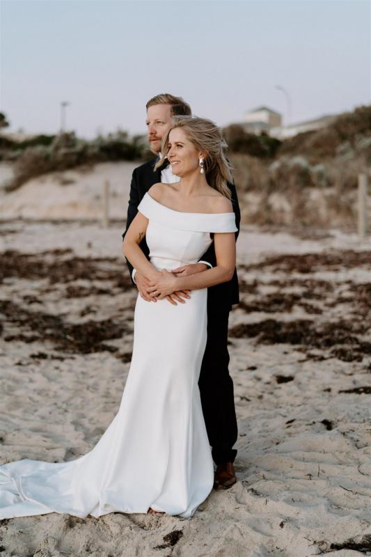 KWH real bride Shelley stands with Jack on a Perth beach. She wears the timeless Lauren wedding dress with long train and off the shoulder sleeve.