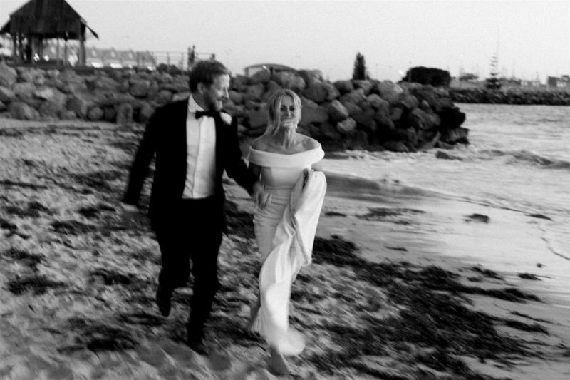 B&W image of KWH real bride Shelley with Jack on the beach. She holds up the long train of her Lauren wedding dress.