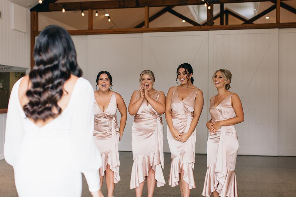 KWH real bride Rebekah's bridesmaids in light pink silk dresses are surprised with their first look at the bride. She wears the long sleeve, v-neck Nikki wedding dress