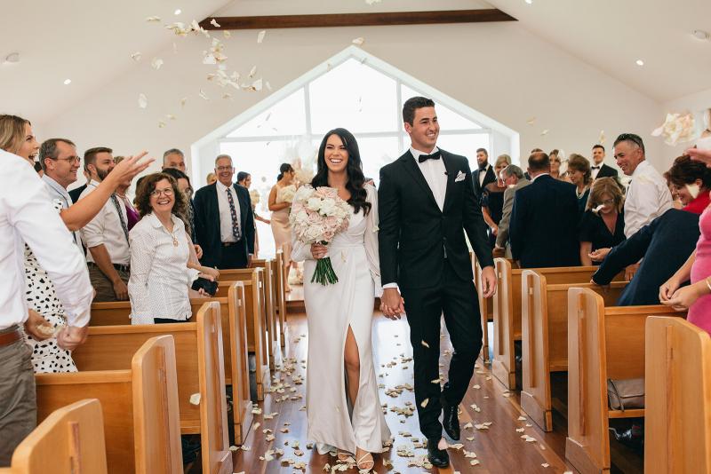 KWH real bride Rebekah walk down the aisle as the rose pedals are thrown in the air. She wears the effortless Nikki wedding dress with leg split.