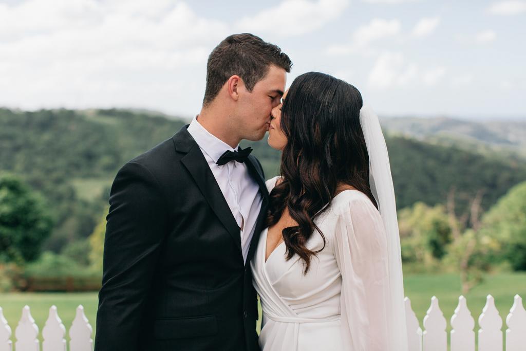 KWH real bride Rebekah kisses her husband outside in her sheer long sleeve Nikki wedding dress.