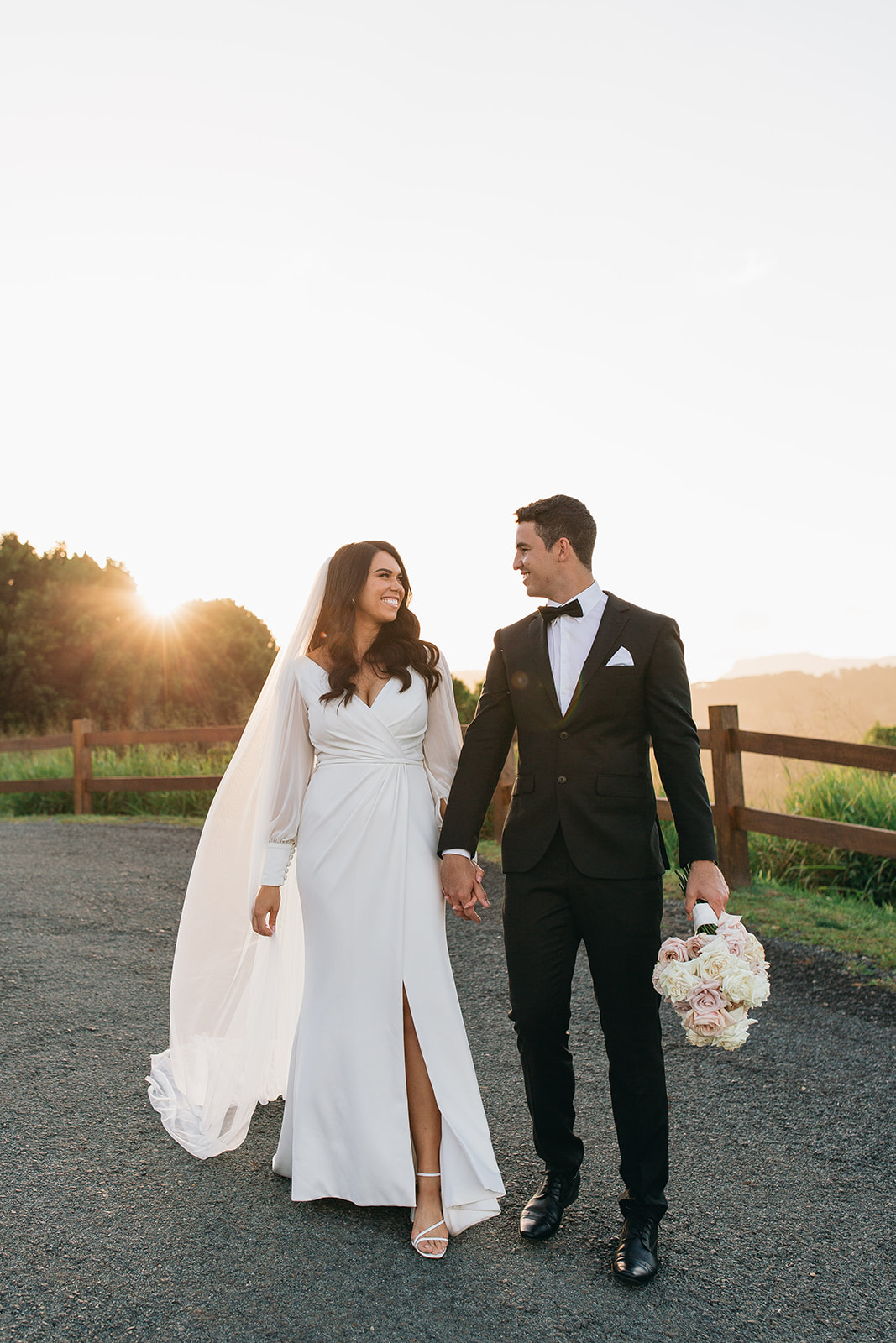 KWH real bride Rebekah and Joel hold hands by the vinyard. She wears the simple Nikki wedding dress with high leg split and long sheer sleeves.