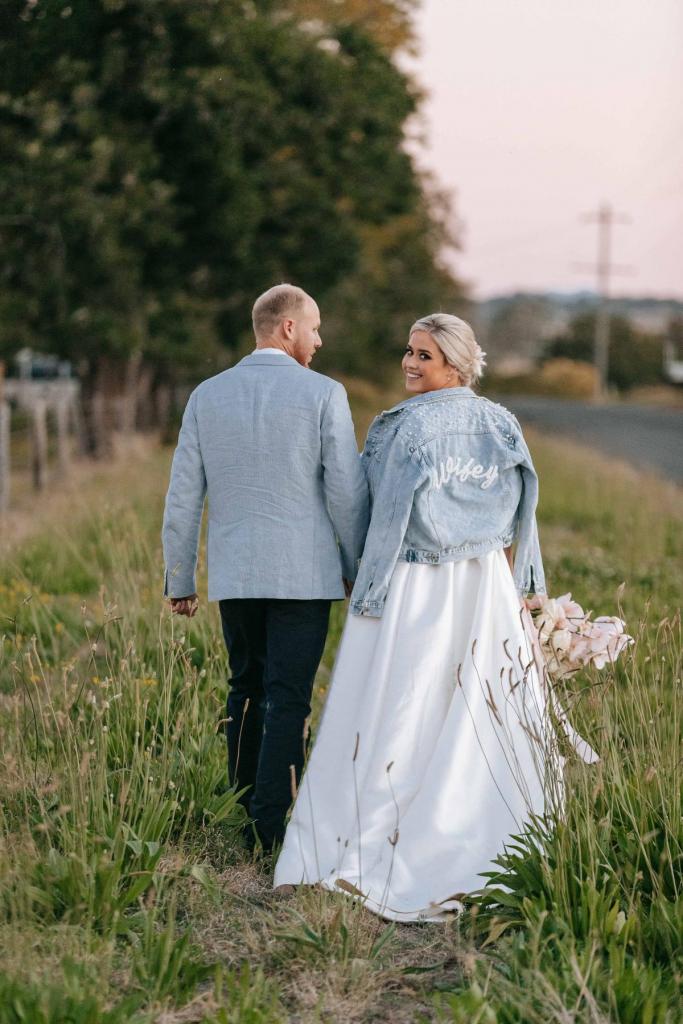 KWH real bride Izzie walking with Jake with her "wifey" embroidered jean jacket. She wears the classic Taryn Camille wedding dress with puff sleeves.