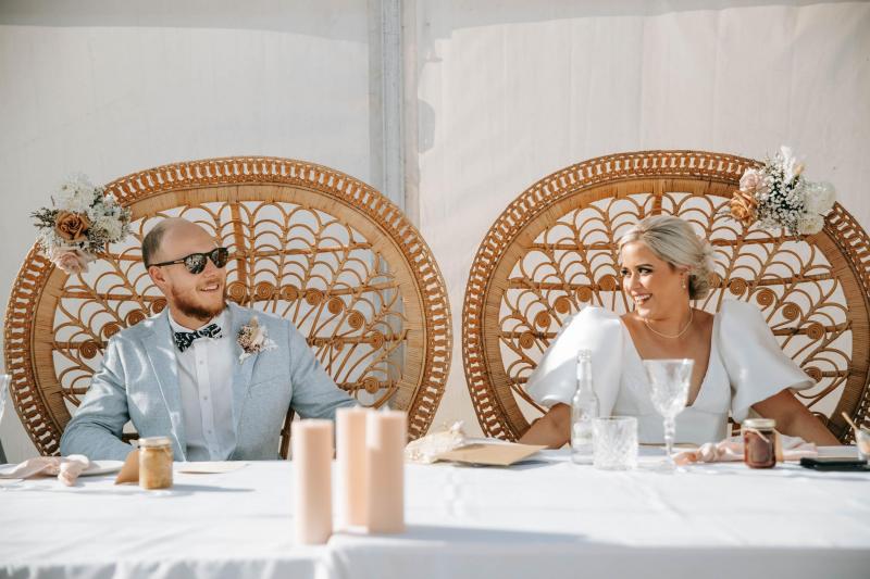 KWH real bride Izzy and new husband Jake sit on rattan chairs for their reception. She wears the Taryn Camille wedding dress with U-shape neckline and bubble sleeves.
