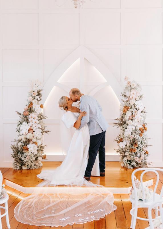 KWH real bride Izzy and Jake kiss at their white chapel alter with floral arches. She wears the classic Taryn Camille with aline skirt and fitted bodice.