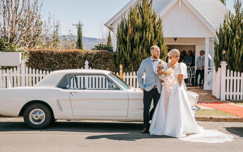 KWH real bride Izzy and Jake stand by a white classic car. She wears the simple Taryn Camille wedding dress with bubble sleeves.