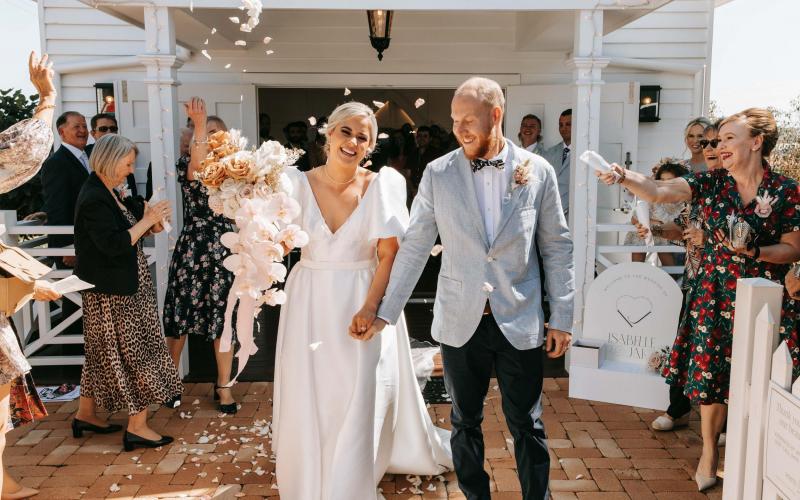 KWH real bride Izzy and Jake walking donw the aisle with petals thrown in the air. She wears the BESPOKE Taryn Camille wedding dress with U-shaped neckline.