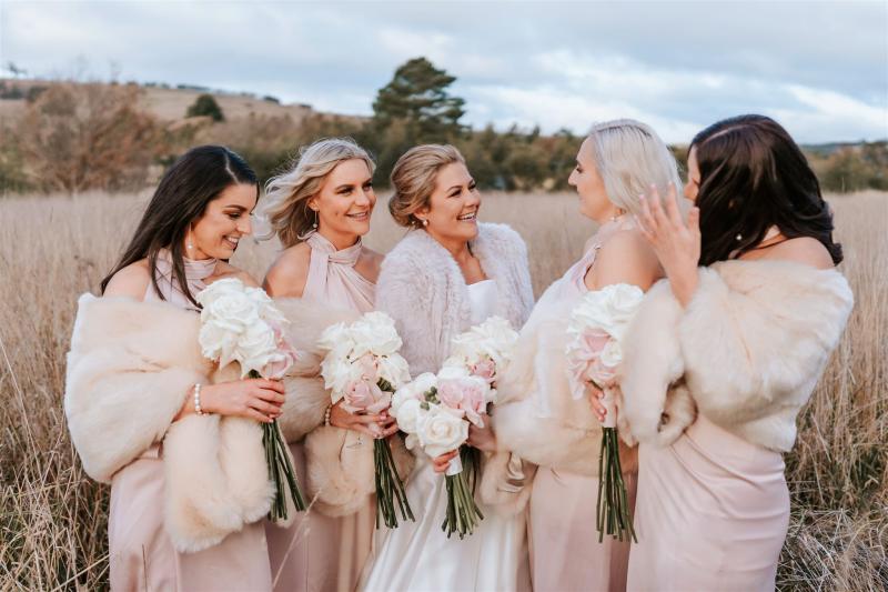 KWH real bride Annabelle standing in a field with her bridesmaids. She wears the classic Jacqueline Melanie gown, a strapless ballgown wedding dress with fur cape.