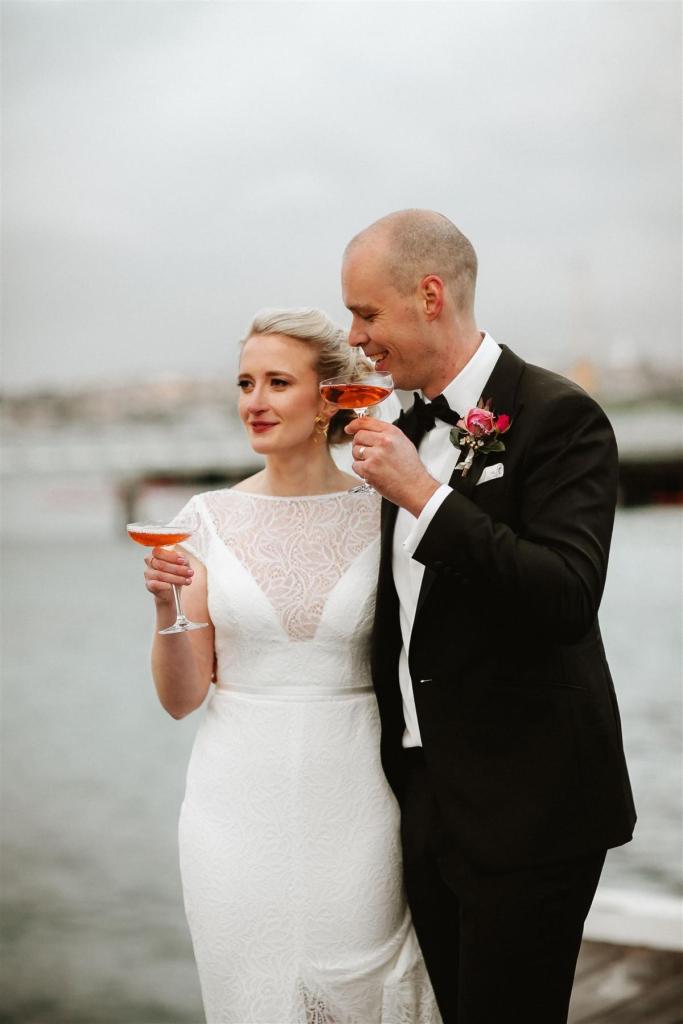 KWH real bride Gemma and Rob stand on a pier holding a cocktail. She wears the Jemma gown, a high neck lace wedding dress with open back.