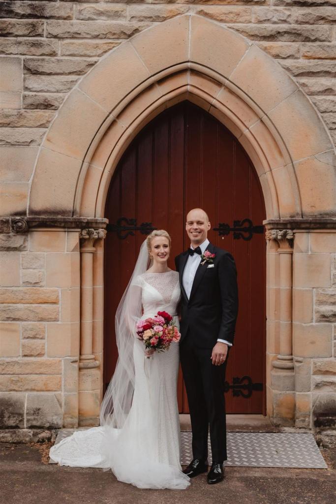 KWH real bride Gemma and Rob standing in front of the church doors. She wears the ivory Jemma gown, a modern high neck lace wedding dress.