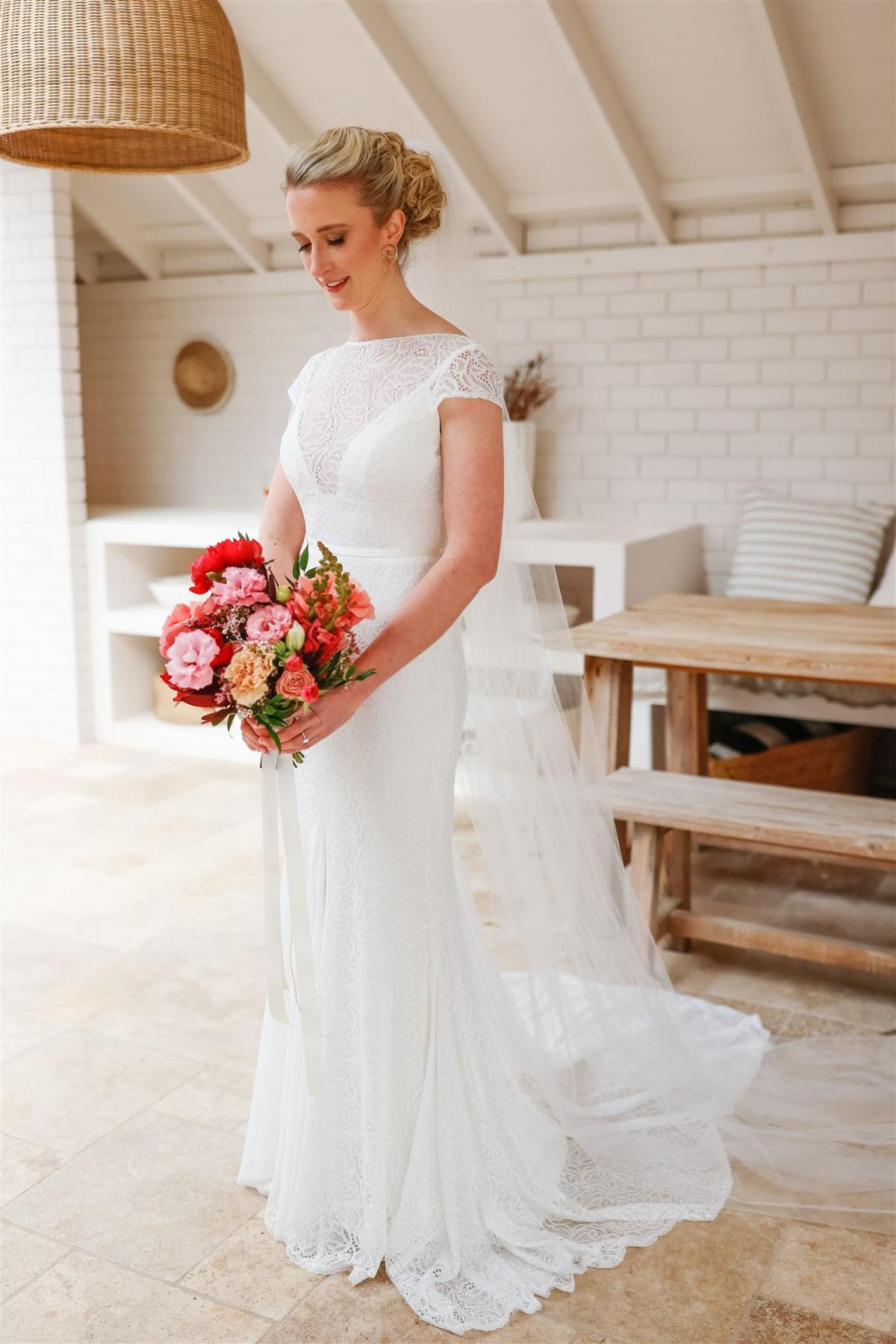 KWH real bride Gemma stands with her bright pink and red bridal bouquet in her Jemma gown, a lace fit and flare high neck wedding dress.