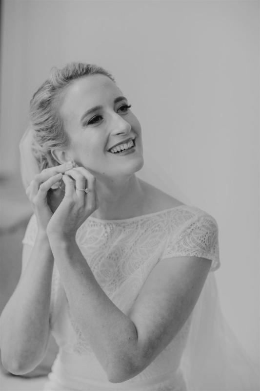 B&W image of Gemma putting in her earring. She wears the modern Jemma gown, a lace fit and flare cap sleeve wedding dress.