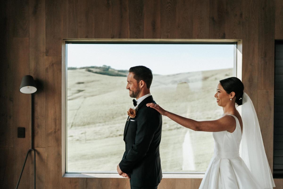 KWH real bride Rebecca taps Matthew's shoulder for their first look as she wears the grand Leonie Melanie gown, a custom v-neck wedding dress with a-line skirt.