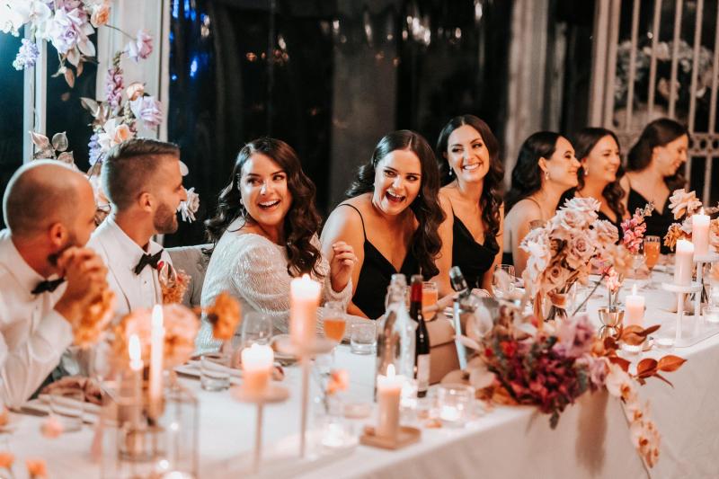KWH real bride Katherine laughs with her bridal party at their wedding table. She wears the modern Margareta gown, a backless beaded wedding dress.