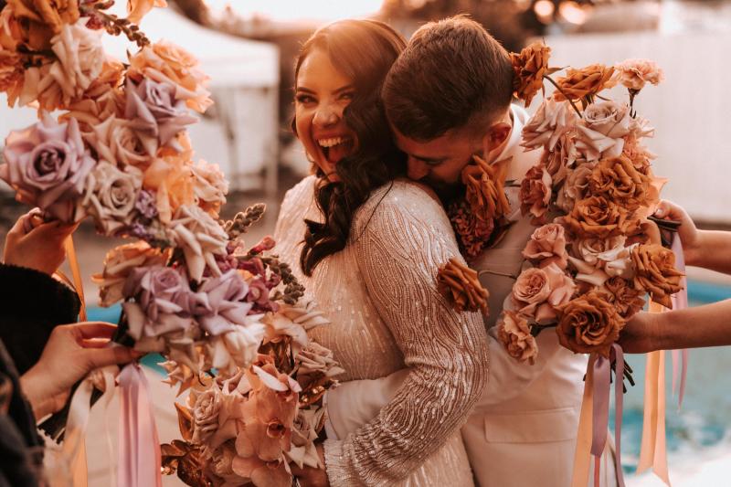 KWH real bride Katherine being snuggled by Conor surrounded by brown and pink roses. She wears the Margareta gown, a beaded fit and flare wedding dress