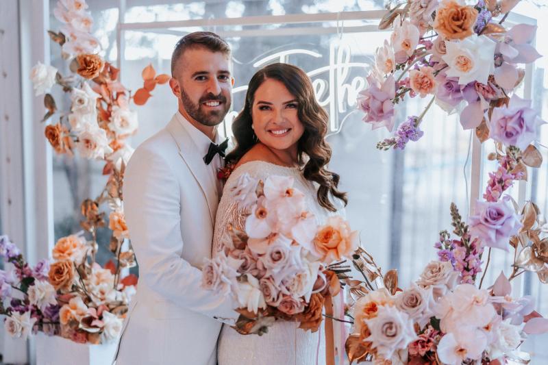 KWH real bride Katherine and Conor smile surrounded by their wedding florals. She wears the glamorous Margareta gown, a high neck fit and flare wedding dress.