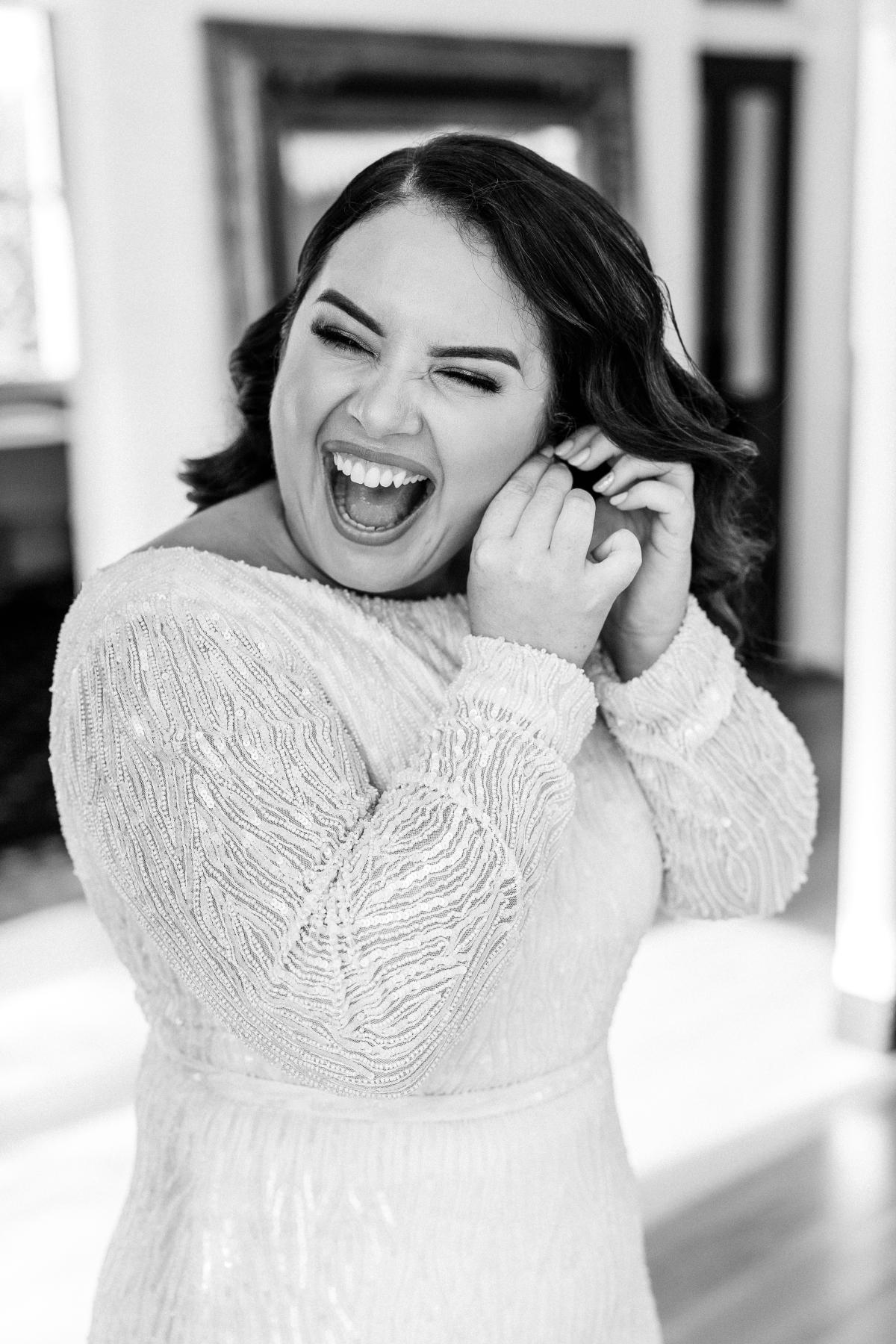 KWH real bride Katherine beams as she puts in her earrings. She wears the glam Margareta gown, a long sleeve beaded wedding dress.