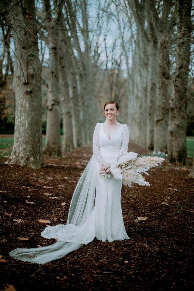KWH real bride Rachel stands in the woods holding her dried flower bouquet in her Rylie gown, a fitted lace wedding dress.