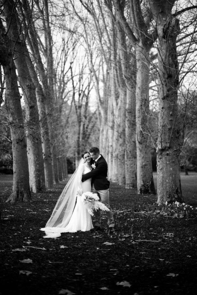 B&W image of KWH real bride Rachel and Josh in the woods. She wears the ivory Rylie gown, a lace fit and flare wedding dress.