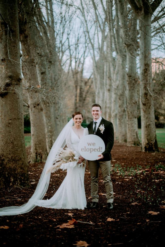 KWH Real bride Rachel and Josh hold up an "elopement" sign. She wears the Rylie gown, a long lace wedding dress with long sleeves.