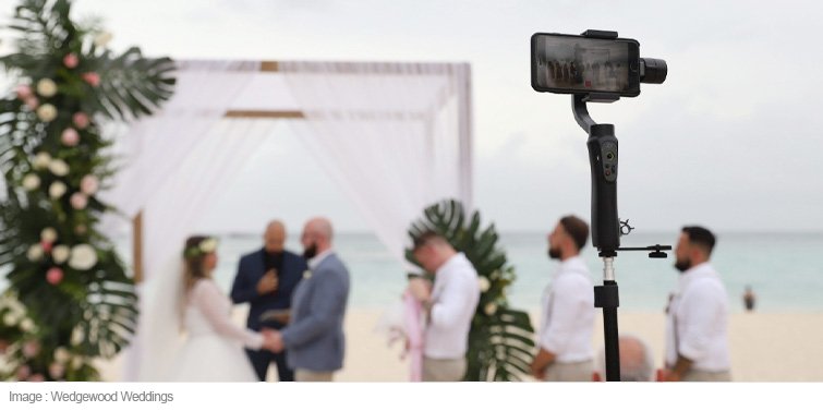 A camera set up on a tripod takes a live stream of a beach wedding.