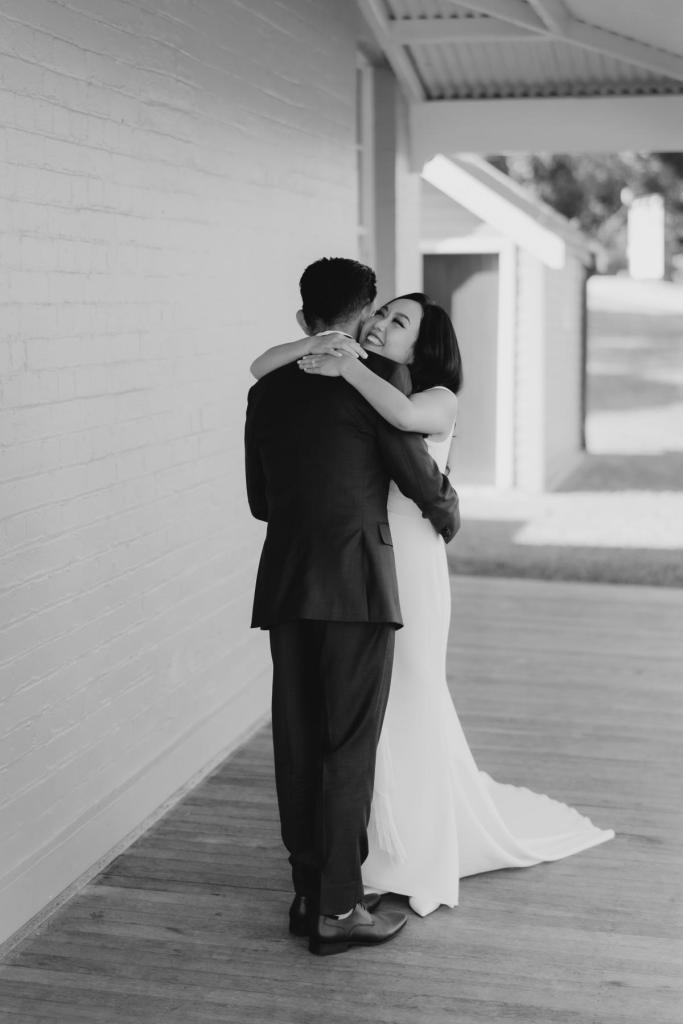 B&W image of KWH real bride Vicki and Tom hugging after their first look. She wears the Arabella gown, plunging neckline simple sheath wedding dress..