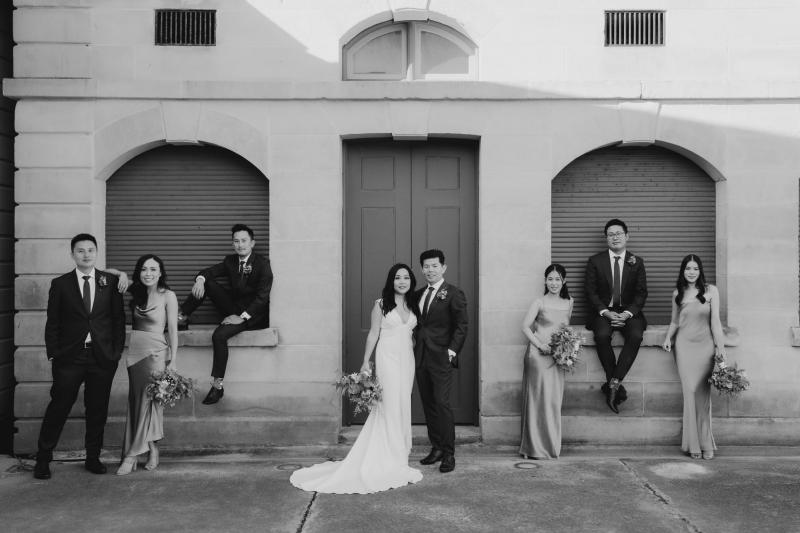 B&W image of KWH real bride Vicki and Tom with their wedding party in front of an old building. She wears the Arabella gown, lunging neckline simple wedding dress.