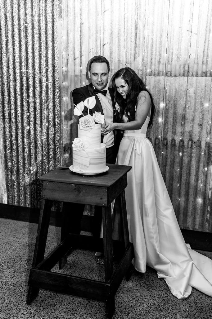 B&W image of KWH real bride Hannah and Reece cutting their cake. She wears the Blake Camille gown, a modern strapless aline wedding dress with leg split..